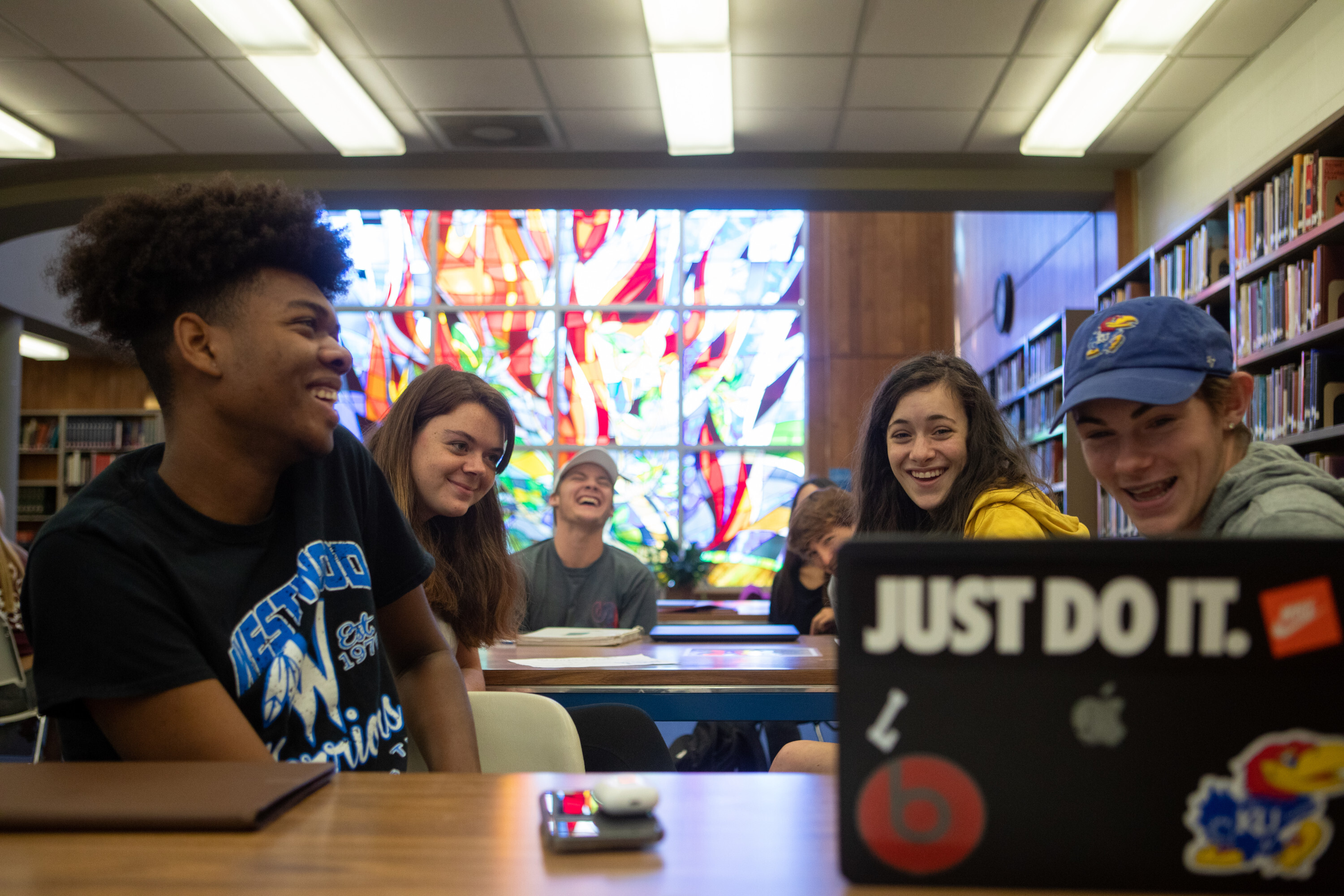 Students seated in a library laughing as they look at a laptop screen